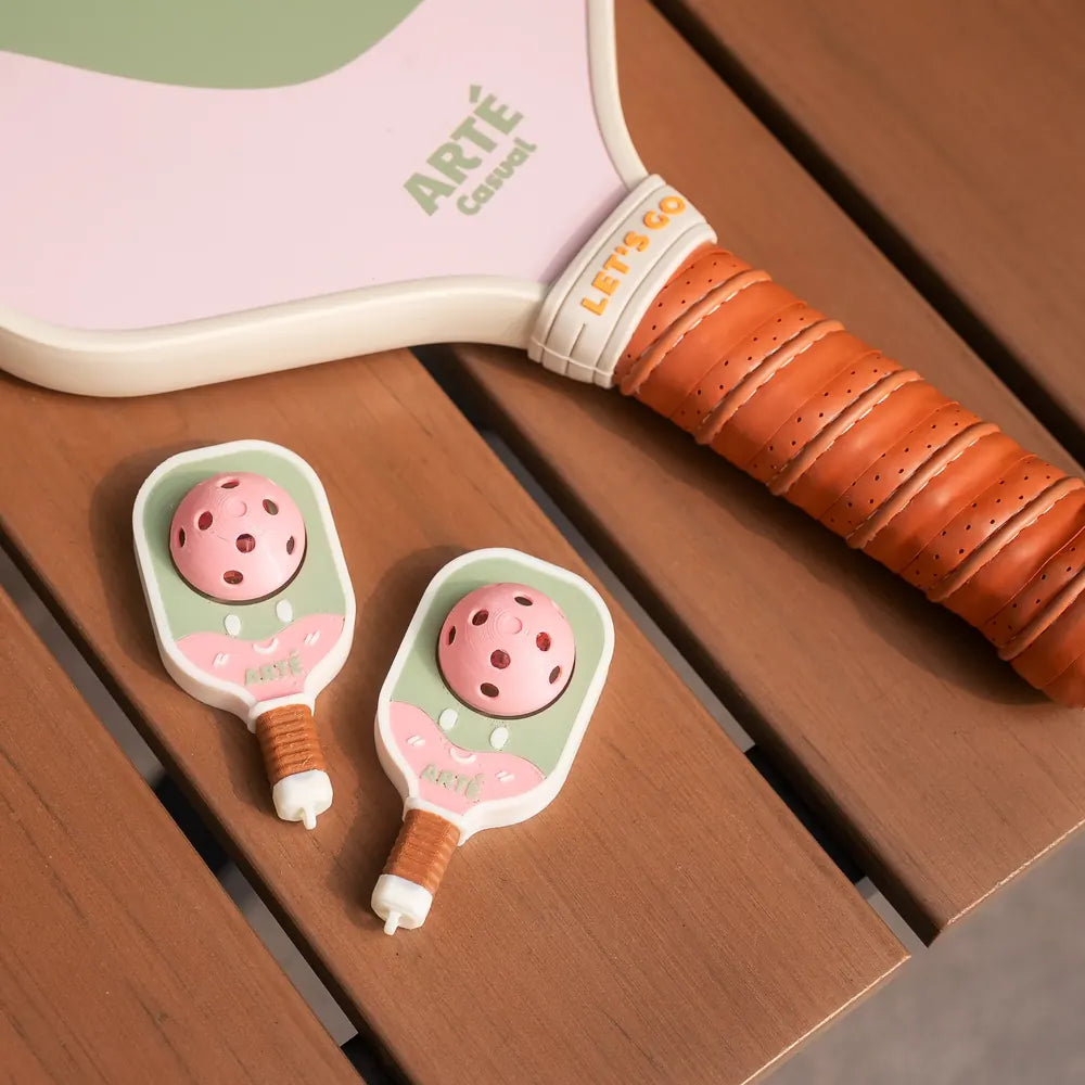 Two Bubbly Clicker charms on a wooden bench beside an ARTÉ Casual paddle in the same green and pink. Paddle not included.